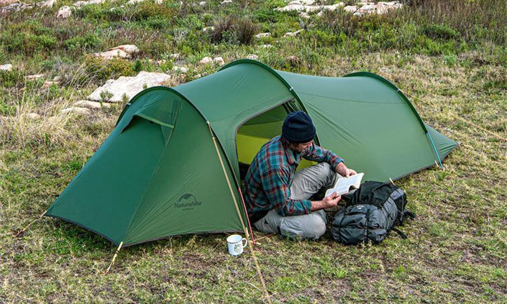 Vihreä Naturehike teltta nurmikolla, mies lukemassa kirjaa teltan edustalla